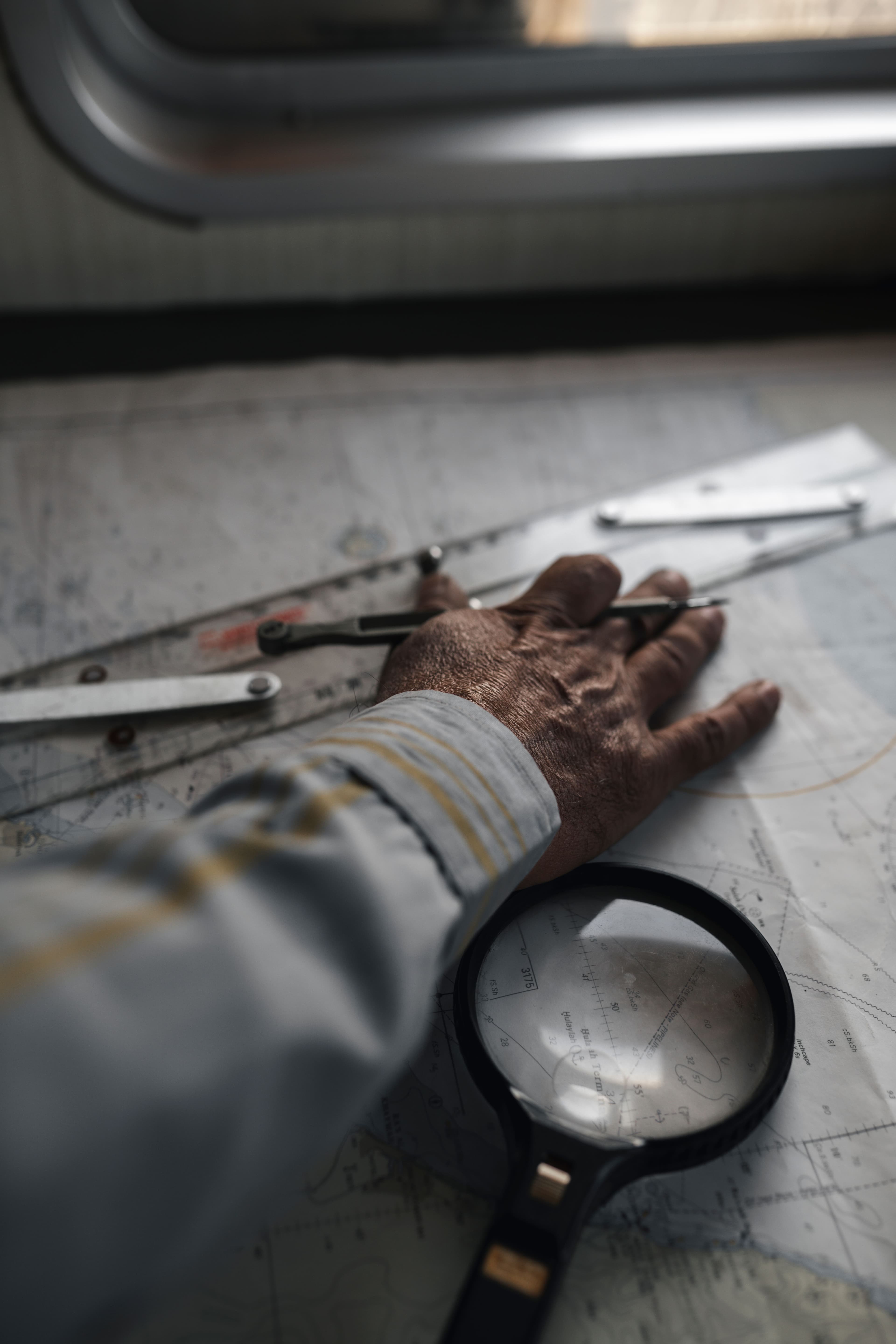 A hand resting on a paper chart beside a magnifying glass and drawing instruments, checking the details